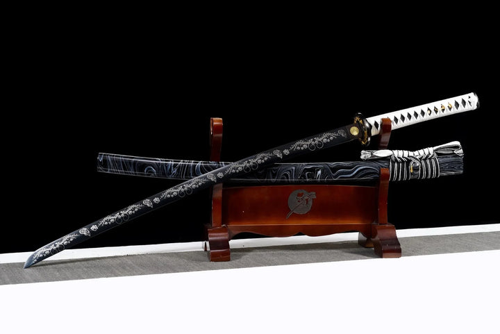 Decorative katana with a black engraved blade and white-wrapped handle, displayed on a wooden stand with its patterned scabbard against a black background.