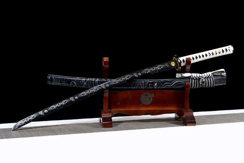 Decorative katana with a black engraved blade and white-wrapped handle, displayed on a wooden stand with its patterned scabbard against a black background.