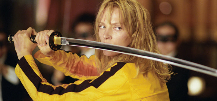 The Bride (Uma Thurman) in a yellow jumpsuit holding a katana sword in a combat stance during a scene from Kill Bill.