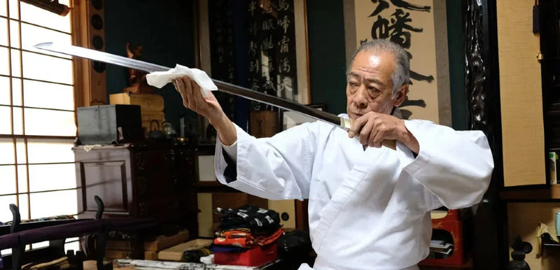 An experienced Japanese swordsman in a white gi carefully inspects the blade of a traditional katana in a classic Japanese dojo setting, with calligraphy scrolls and sword-polishing cloth in hand.