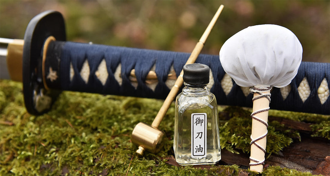 Katana maintenance kit displayed on moss, featuring a sheathed sword, oil bottle, brass hammer, and powder ball for blade care.