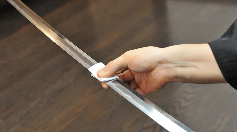 A hand using a small white cloth to carefully polish and clean the flat of a katana blade resting on a wooden surface.