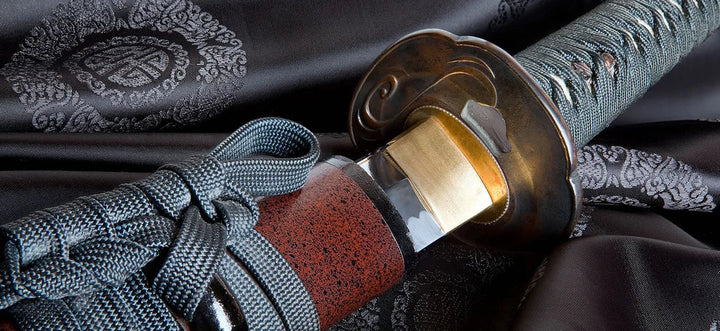 Close-up of a katana hilt and guard resting on patterned fabric, showing detailed wrap, cord, and metal fittings.
