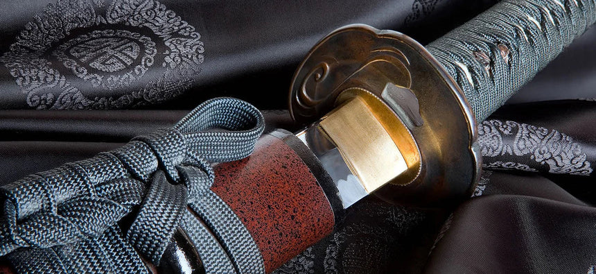 Close-up of a katana hilt and guard resting on patterned fabric, showing detailed wrap, cord, and metal fittings.