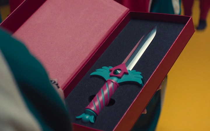 A decorative dagger with a pink and teal ornate handle displayed in a red presentation box, appearing to be from Squid Game Season 2.