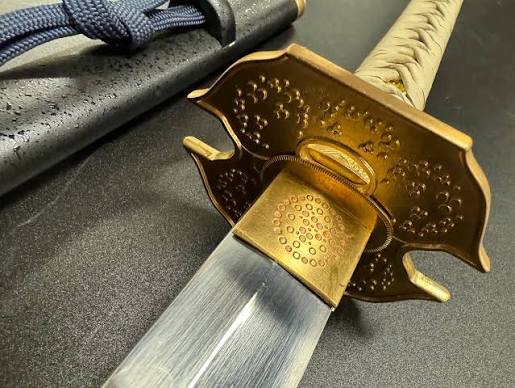 Close-up of a katana's gold-toned guard and polished blade, with a wrapped handle and black scabbard visible in the background.