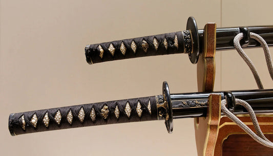 Two traditional Japanese katana swords resting on a wooden display stand, featuring black wrapped handles with white diamond-pattern inlays and decorative metal fittings.