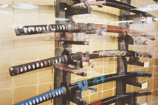 Several decorative katana swords displayed horizontally on a wooden rack behind glass, featuring curved blades, wrapped handles, and ornate scabbards.
